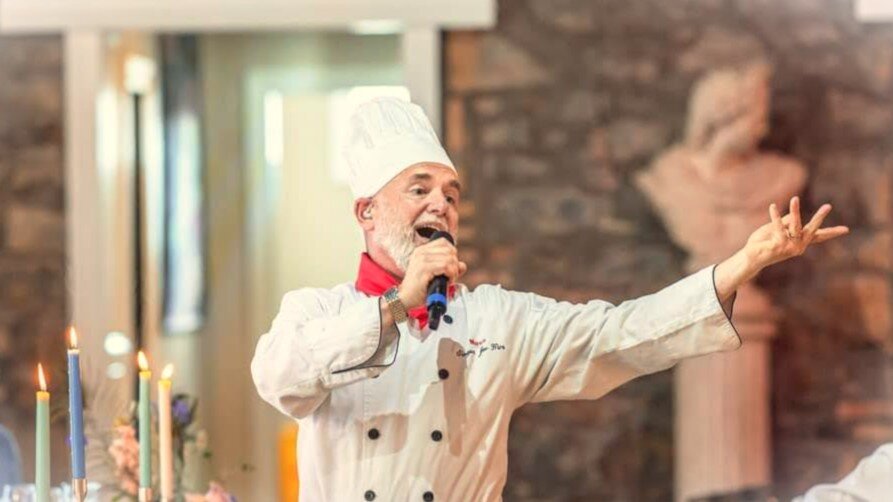 Chef in full uniform singing into a microphone during a lively performance with candles and décor in the background.