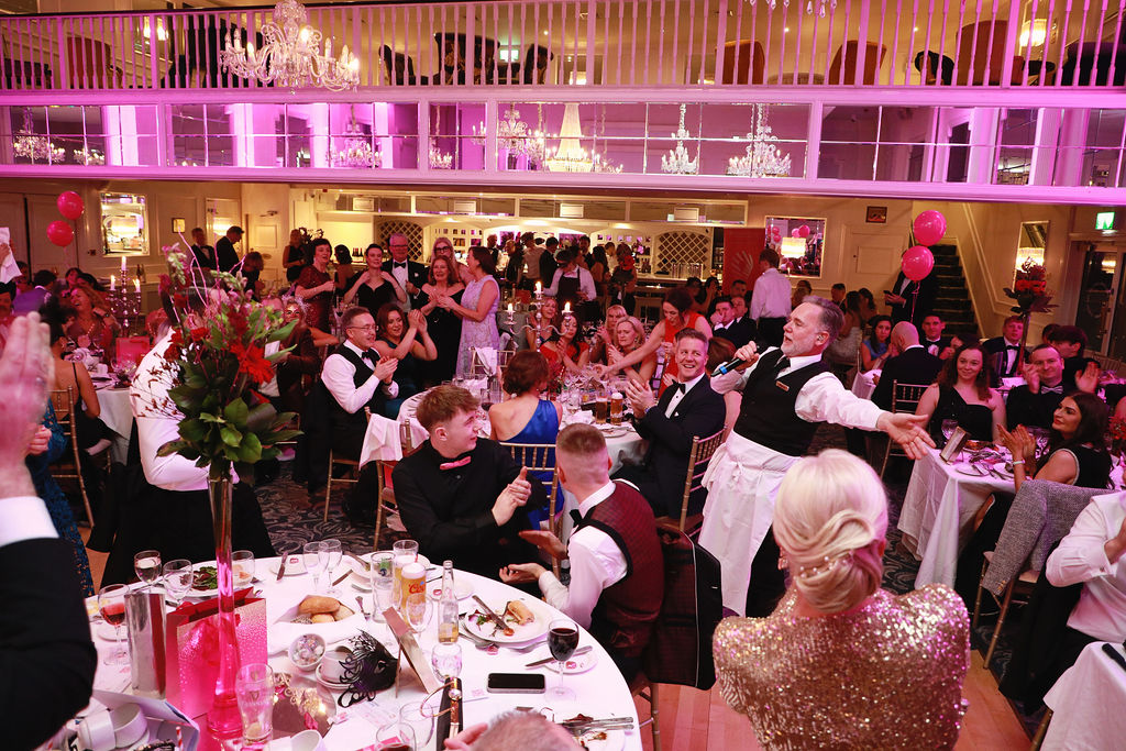“Singing waiter addressing an enthusiastic audience at a formal dinner, with guests raising their hands in response.”
