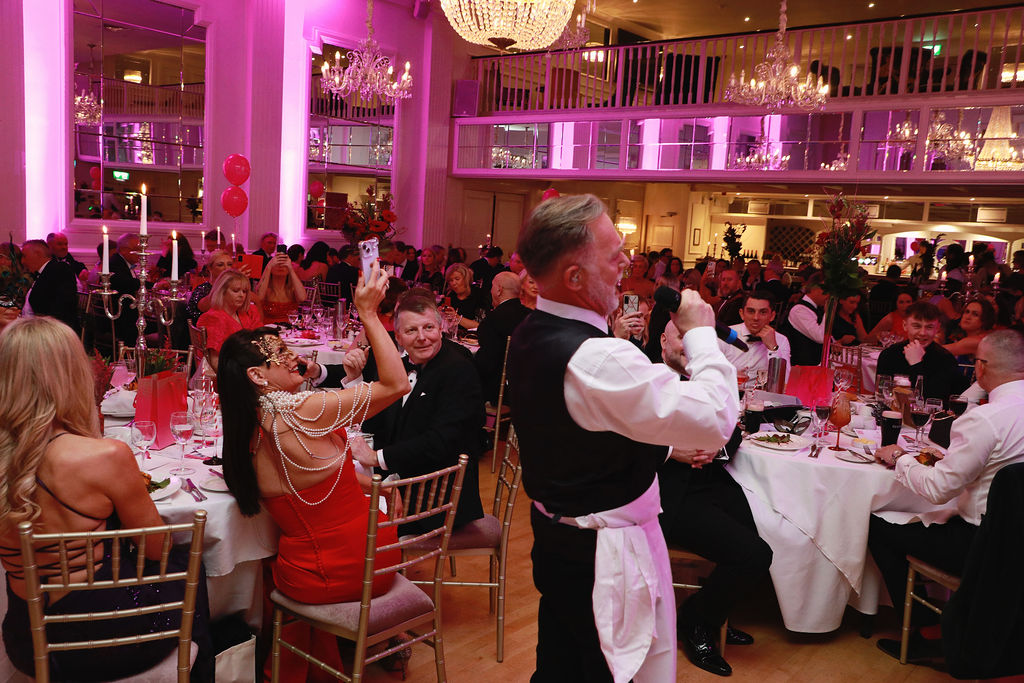 “Professional singing waiter performing among guests at a formal dinner, with attendees recording the moment on their phones.”