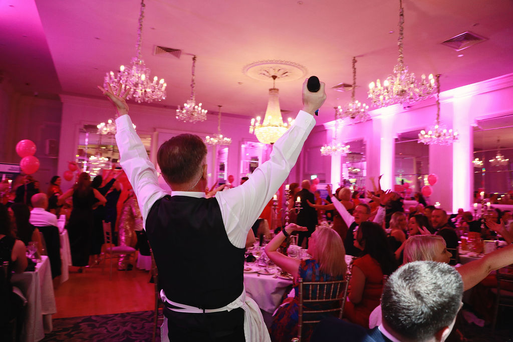 “Professional singing waiter performing with a microphone at a formal gala dinner, entertaining guests seated at elegantly decorated tables.”